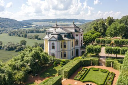 Rokokoschloss mit pastellfarbener Fassade, umgeben von symmetrischem Schlossgarten und grüner Landschaft bei Sonnenschein