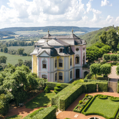 Rokokoschloss mit pastellfarbener Fassade, umgeben von symmetrischem Schlossgarten und grüner Landschaft bei Sonnenschein