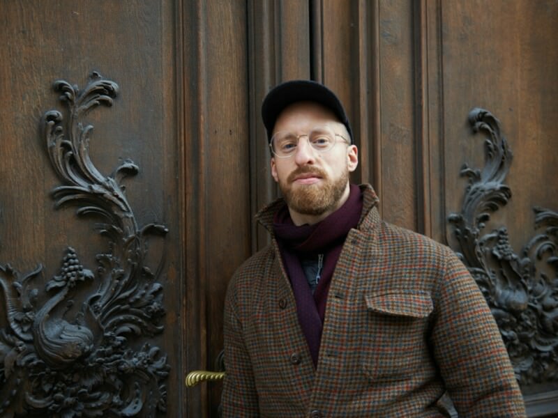 Der Influencer Baroqueblockbuster mit Brille, Bart und Mütze steht vor einer kunstvoll verzierten Holztür. Die Tür hat aufwändige Schnitzereien im barocken Stil, die Pflanzenmotive und filigrane Muster zeigen. Der Mann trägt einen karierten Mantel, einen Schal und eine schwarze Mütze und blickt ruhig in die Kamera.
