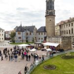 Mehrere Menschen versammeln sich tagsüber auf einem Platz vor dem Weimarer Stadtschloss zu einer Open-Air-Veranstaltung. Es gibt eine kleine Bühne mit weißen Sonnenschirmen und Sitzgelegenheiten. Im Hintergrund sind der Bastille-Turm, historische Gebäude und Teile eines Gartens mit Blumenbeeten zu sehen.