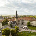 Luftaufnahme des Weimarer Stadtschlosses mit Bastille-Turm bei Abendlicht. Vor dem Schloss findet eine Veranstaltung mit vielen Menschen, Sitzplätzen und weißen Pavillons statt. Im Vordergrund sitzen Besucher auf Picknickdecken im Schlossgarten. Im Hintergrund sind die Stadt Weimar mit Häusern, Kirchtürmen und Bäumen zu sehen.