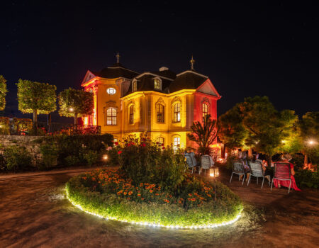 Illuminiertes Rokokoschloss Dornburg bei Nacht, mit festlich beleuchtetem Garten, Besucherinnen und Besuchern auf Stühlen unter Bäumen – Aufnahme während der Dornburger Schlössernacht im Sommer in Thüringen.